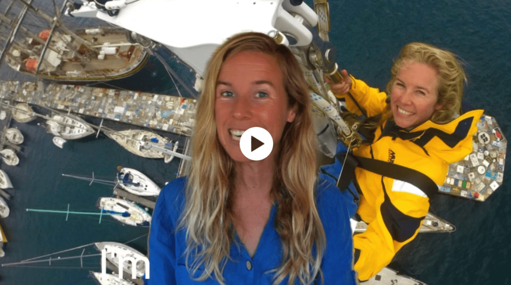 A woman in a yellow jacket is standing next to a group of sailboats, surrounded by a sailing crew.