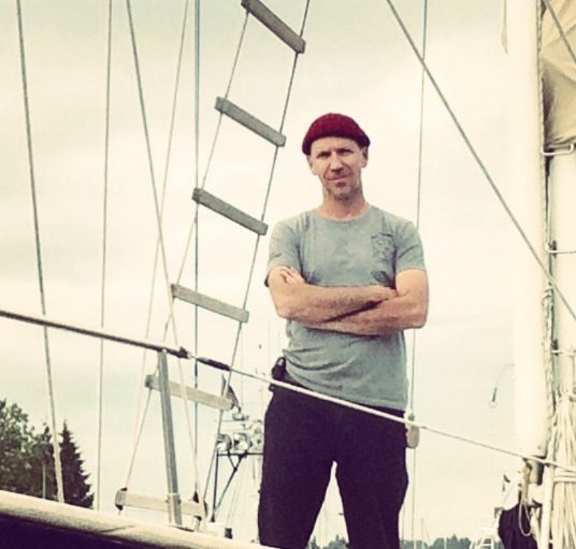 A person in a red hat and gray shirt stands with crossed arms on a boat deck, embodying the spirit of the Ocean Nomads, with ropes and a ladder visible in the background.