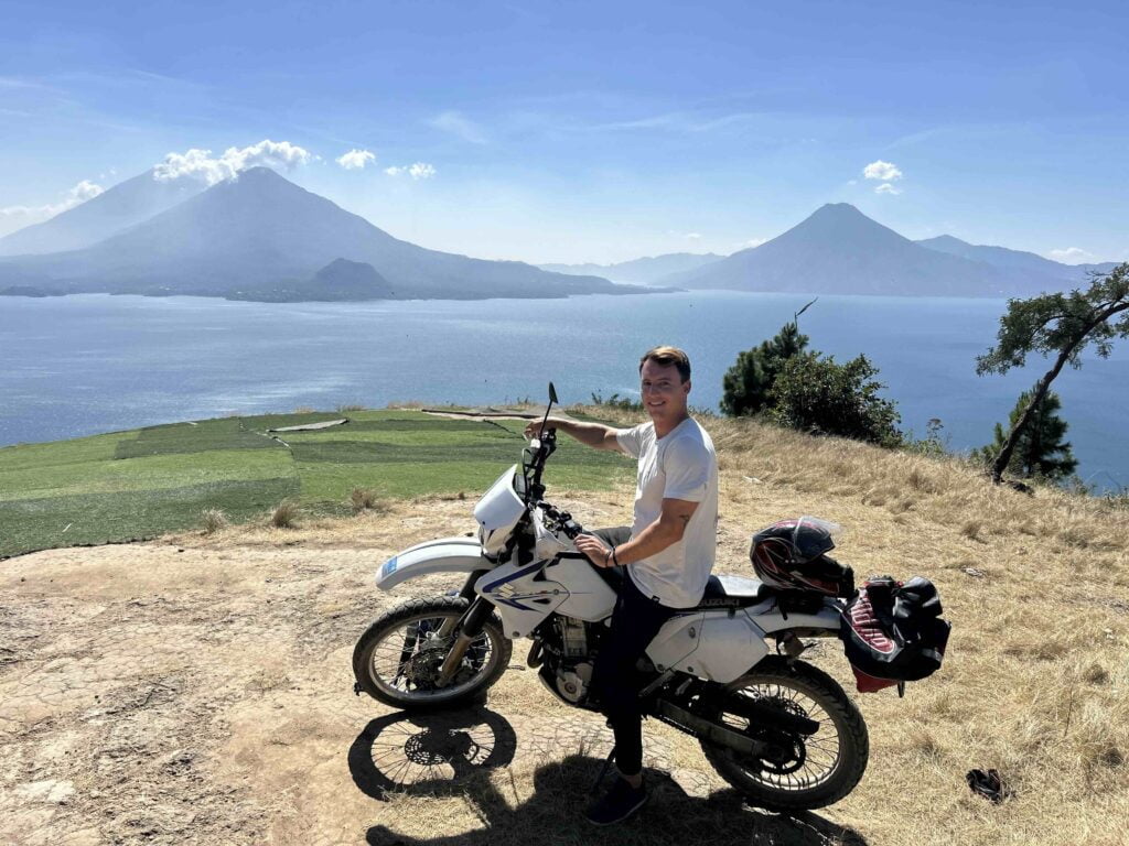 A world sailor sits on a motorcycle near a grassy cliff with a scenic view of a lake and distant mountains in the background.