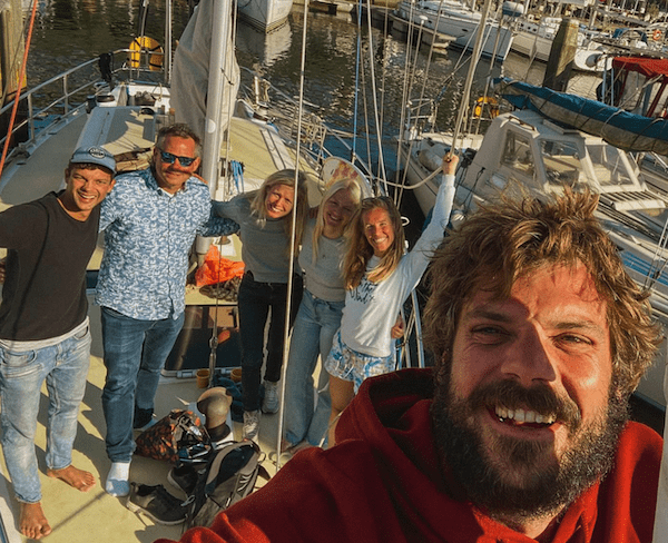A group of five people, including a marine biologist, stand on a boat, smiling at the camera with water and other boats in the background. One person in a red hoodie is taking the selfie.