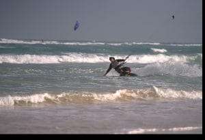 A person kite surfing near the shore, leaning back on the board with waves and another kite visible in the background.